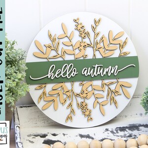 May include: A round, white wooden sign with laser-cut botanical elements and the words "hello autumn." The sign features a green band across the center with the greeting in white script. The design includes beige-colored leaves and floral accents.