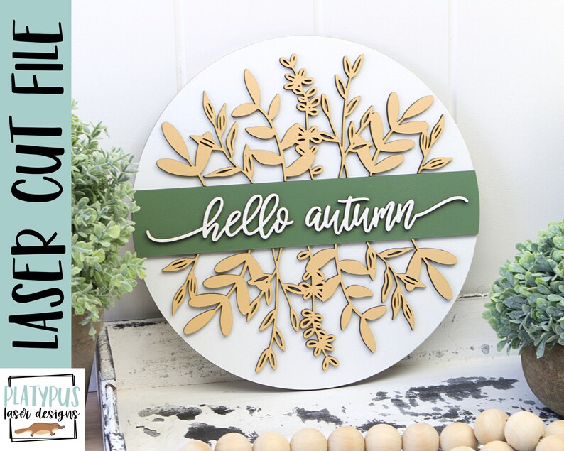 May include: A round, white wooden sign with laser-cut botanical elements and the words "hello autumn." The sign features a green band across the center with the greeting in white script. The design includes beige-colored leaves and floral accents.