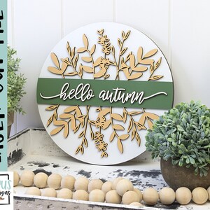 May include: A round wooden sign with a white background and laser-cut botanical design in beige. A green band across the center reads "hello autumn" in white script. The sign is a laser cut file.
