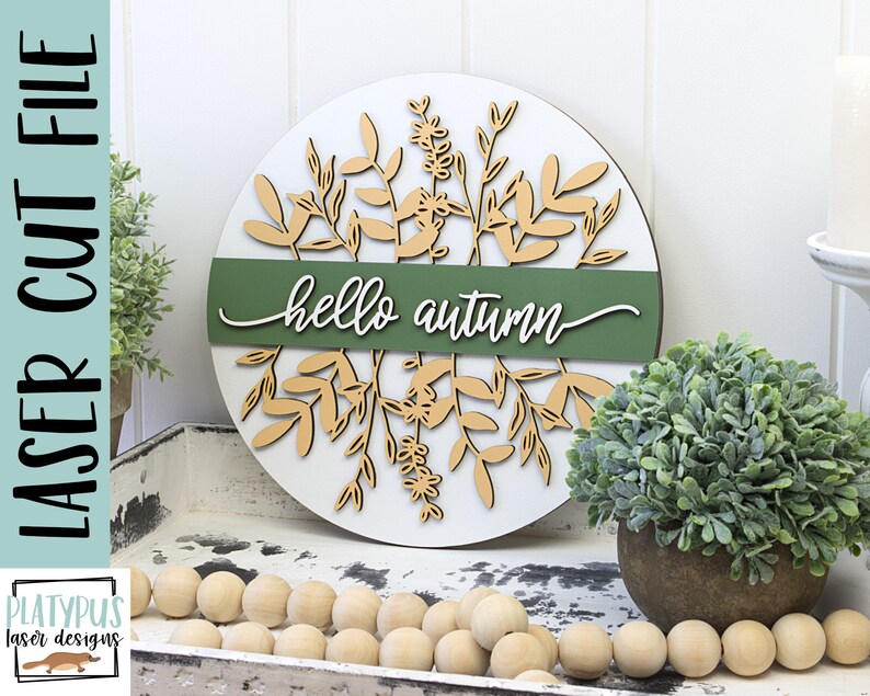 May include: A round wooden sign with a white background and laser-cut botanical design in beige. A green band across the center reads "hello autumn" in white script. The sign is a laser cut file.
