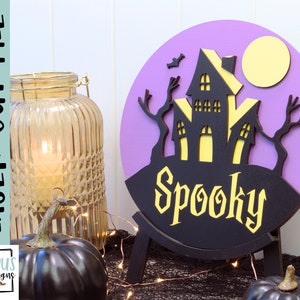 May include: A purple and black layered wooden Halloween sign with a spooky house, trees, and a moon. The sign says "Spooky" in yellow letters.