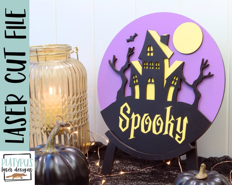 May include: A purple and black layered wooden Halloween sign with a spooky house, trees, and a moon. The sign says "Spooky" in yellow letters.