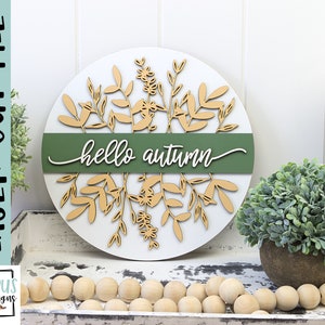 May include: A round, white wooden sign with laser-cut botanical elements and the words "hello autumn" in white script. A green band bisects the sign. The sign is decorated with light brown leaves and floral designs.