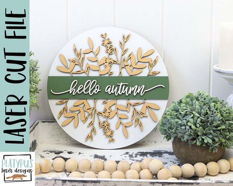 May include: A round, white wooden sign with laser-cut botanical elements and the words "hello autumn" in white script. A green band bisects the sign. The sign is decorated with light brown leaves and floral designs.
