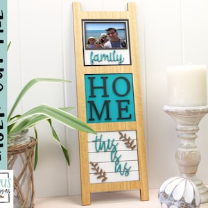 May include: Wooden picture frame with a photo of a family, the word "family" in cursive, the word "HOME" in black letters on a teal background, and the words "this is us" in white letters on a wood background.