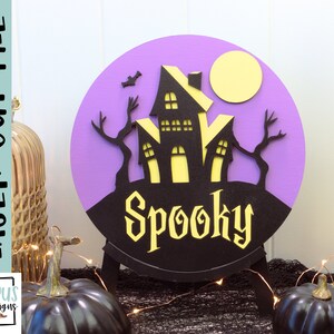 May include: A purple and black round wooden sign with a spooky haunted house silhouette, a yellow moon, and the word "Spooky" in a black and yellow font.