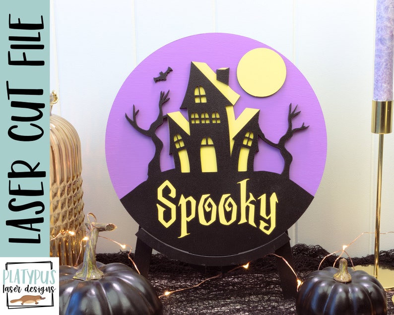 May include: A purple and black round wooden sign with a spooky haunted house silhouette, a yellow moon, and the word "Spooky" in a black and yellow font.