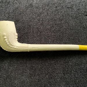 Puede incluir: Una pipa de tabaco de color crema con un cuenco decorativo y un largo tallo. El cuenco presenta un diseño ornamentado con detalles redondeados. El tallo está pintado de color crema, con una punta amarilla. La pipa descansa sobre una superficie gris oscuro.