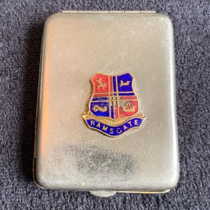 Puede incluir: Una cajetilla de cigarrillos de metal plateado con un escudo de esmalte de colores que presenta un escudo con un caballo rojo, un barco azul y un ciervo. El escudo está rodeado por un borde dorado y el texto "RAMSGATE" en letras doradas.
