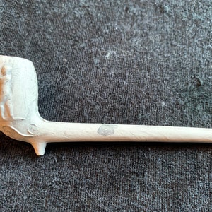 May include: A white ceramic pipe with a carved design of a person on the bowl.