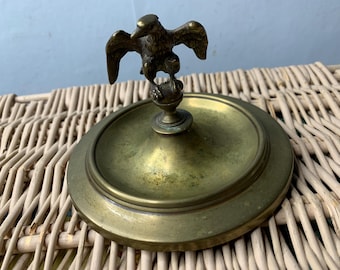 May include: A brass trinket dish with a raised eagle design on the lid. The dish is round and has a slightly raised rim.