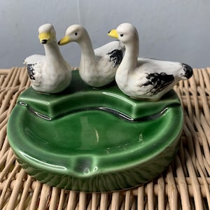 May include: A green ceramic ashtray with three white ceramic geese with black markings and yellow beaks. The geese are perched on the edge of the ashtray.