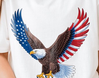 May include: White t-shirt featuring a graphic of a bald eagle with wings spread. The eagle's wings are colored with the American flag design, with blue and white stars on one wing and red and white stripes on the other. The eagle has a brown body and yellow talons.