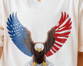 May include: Cream-colored t-shirt featuring a detailed graphic of an eagle with wings spread wide. The eagle's wings are colored with the American flag design, with blue and white stars on one side and red and white stripes on the other.