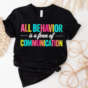 May include: Black t-shirt with the text "ALL BEHAVIOR is a form of COMMUNICATION" in bright, colorful letters. The text is in yellow, teal, orange, pink, and green. The shirt is folded at the bottom.