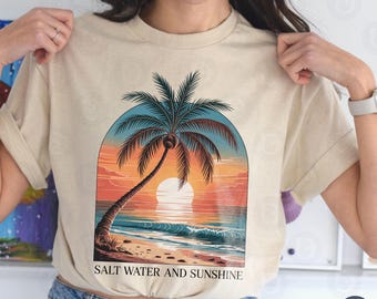 May include: Beige t-shirt featuring a tropical sunset scene with a palm tree, ocean waves, and a setting sun. The text "SALT WATER AND SUNSHINE" is printed at the bottom of the graphic. The shirt has short sleeves and a relaxed fit.