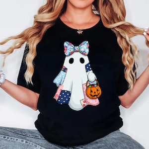 May include: Black t-shirt featuring a cartoon ghost with a floral bow and patchwork design. The ghost holds a jack-o'-lantern bucket, perfect for Halloween. The design incorporates blue, pink, and white colors.
