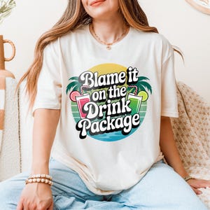 Könnte beinhalten: Cremefarbenes T-Shirt mit dem Text "Blame it on the Drink Package" in Retro-Design. Die Grafik zeigt Palmen, einen Sonnenuntergang und Cocktail-Bilder. Das Shirt wird mit einer Jeans getragen.