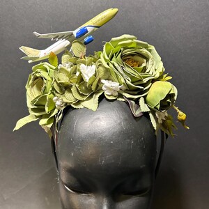 May include: A green floral headband with a small airplane charm. The headband is made of artificial flowers and leaves.