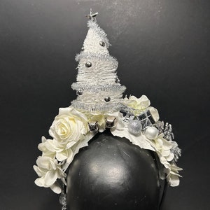 Può includere: Un cerchietto per capelli natalizio bianco con un mini albero di Natale bianco floccato, fiori bianchi, campane d'argento e ornamenti d'argento.