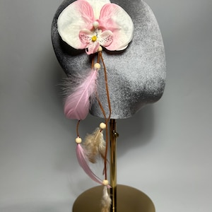 Può includere: Copricapo decorativo con una grande orchidea bianca e rosa. Piume rosa e perline di legno pendono dal fiore, attaccate con uno spago marrone. Il copricapo è esposto su una forma di velluto grigio, montata su un supporto dorato.