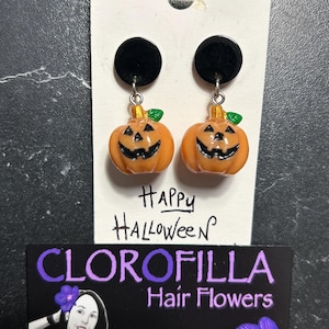 Può includere: Orecchini a tema Halloween con ciondoli a forma di zucca arancione con facce sorridenti e foglie verdi. Gli orecchini pendono da borchie circolari nere. Il testo "Happy Halloween" è visibile.