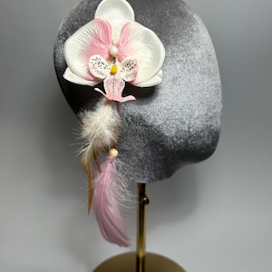 Può includere: Un copricapo in velluto grigio ornato da un fiore di orchidea bianco e rosa, accentuato da piume. L'orchidea ha un centro giallo e delicati accenti rosa. Le piume sono un mix di bianco, marrone e rosa.