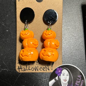Può includere: Un paio di orecchini a tema Halloween. Ogni orecchino presenta tre zucche arancioni impilate con facce intagliate, sospese da un disco nero. Gli orecchini sono attaccati a una carta marrone con le parole "Happy Halloween" e "Pumpkins Totem".