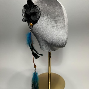 Può includere: Un fermaglio per capelli a forma di fiore nero con dettagli glitterati, ornato da piume blu e nere e piccole perline. L'accessorio per capelli è esposto su una testa di velluto grigio, montata su un supporto dorato.