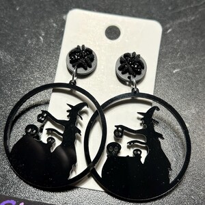 Può includere: Orecchini neri a tema Halloween. Ogni orecchino presenta un design circolare con una silhouette di strega, un calderone e dettagli di teschio. La parte superiore di ogni orecchino ha un piccolo elemento decorativo rotondo nero.