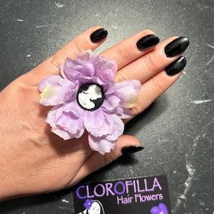 Può includere: Accessorio per capelli a forma di fiore lilla con un cammeo al centro. Il fiore è in tessuto e ha un cammeo bianco e nero al centro. È visibile anche un biglietto da visita con la scritta "CLOROFILLA Hair Flowers".