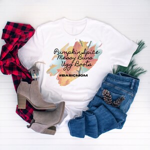 May include: White t-shirt with the text "Pumpkin Spice, Messy Buns, Ugg Boots" and the hashtag "#BASICMOM". The shirt is styled with blue jeans, grey ankle boots, and a red and black plaid scarf.