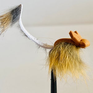 May include: A fly fishing lure with a tan body and a curved white tail with brown and black details. The lure features a small, tan, foam-like head. The fly is designed to resemble an insect and is attached to a black hook.