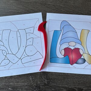 Puede incluir: Dos hojas de papel con un dibujo en blanco y negro del  la palabra "LOVE" con un gnomo sosteniendo un corazón en el medio de la palabra. Las hojas están al lado de una herramienta de plástico roja y una herramienta de plata.