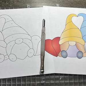 May include: A stained glass pattern for two gnomes holding a heart shape. The pattern is numbered and includes instructions for cutting the glass. The pattern is printed on white paper.