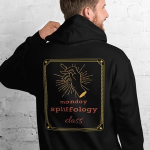 Peut inclure: Sweat-shirt à capuche noir avec un imprimé graphique d'une main tenant un joint. Le graphique est entouré d'un cadre doré et le texte "monday spliffology class" est imprimé sous le graphique.