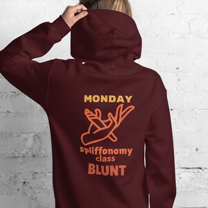 Puede incluir: Sudadera con capucha color burdeos con un gráfico de una mano sosteniendo un porro y el texto "MONDAY spliffonomy class BLUNT".