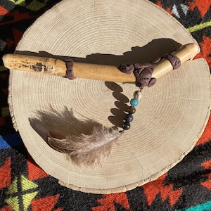 Può includere: Una pipa di legno con dettagli in argilla marrone e una piuma attaccata a una corda di perline con perline nere, marroni e blu.