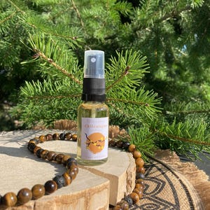 May include: A small spray bottle with a white label that reads "Chileuague" sits on a wooden surface. A beaded necklace with brown and black beads is draped around the bottle. The background features green pine branches.