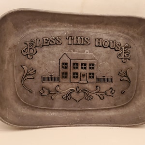 May include: A silver metal tray with the words "Bless This House" embossed on it. The tray features a stylized house with windows and a heart in the door, surrounded by floral designs.