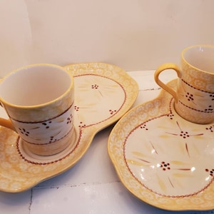 May include: Two yellow ceramic mugs with brown floral designs sit on matching yellow ceramic plates with brown floral designs.