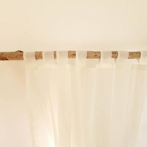 Könnte beinhalten: Transparente, weiße Vorhänge, die an einer Naturholzstange hängen. Die Vorhänge bestehen aus einem leichten, fließenden Stoff und sind mit weißen Stoffschlaufen an der Stange befestigt. Der Hintergrund ist cremefarben.