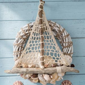Könnte beinhalten: Ein runder Kranz aus weißen geflochtenen Zweigen, verziert mit verschiedenen Muscheln und einer zentralen Treibholzstruktur in Form eines Segelbootmastes mit einem beigen geknoteten Seilnetz. Weitere Muscheln sind darunter verstreut.