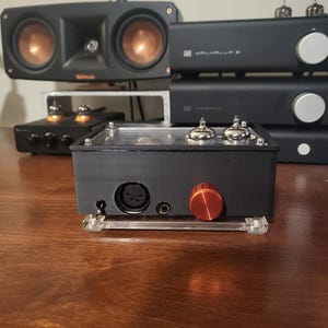 May include: A black audio amplifier with a clear top, silver tubes, and an orange knob. Other black audio components and speakers with copper-colored cones are in the background. The components are on a wooden surface.