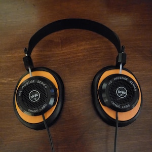 May include: Black and orange Grado SR80e headphones. The headphones have a black headband and earcups with orange accents. The text "THE PRESTIGE SERIES SR 80 e GRADO LABS" is printed on the earcups. The headphones are on a wooden surface.