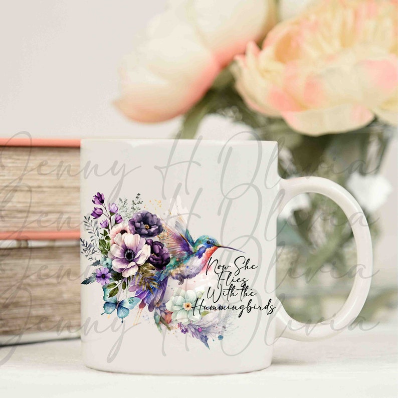 May include: White ceramic mug with a watercolor design of a hummingbird flying with purple and pink flowers. The text "Now She Flies With The Hummingbirds" is printed on the mug.