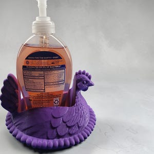 Puede incluir: Un soporte dispensador de jab&oacute;n con forma de gallina morada. El soporte est&aacute; dise&ntilde;ado para sostener una botella transparente de jab&oacute;n de manos antibacteriano. La botella de jab&oacute;n tiene una bomba blanca y l&iacute;quido naranja. El texto "GOOD FOR THE EARTH + BODY" es visible.
