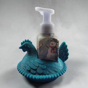 Puede incluir: Un soporte dispensador de jab&oacute;n en forma de gallina de color turquesa con un dise&ntilde;o decorativo. Un dispensador de jab&oacute;n transparente con una bomba blanca se encuentra dentro del soporte. El dispensador de jab&oacute;n tiene una etiqueta con tem&aacute;tica invernal con un mu&ntilde;eco de nieve.