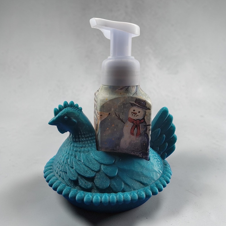 Puede incluir: Un soporte dispensador de jab&oacute;n en forma de gallina de color turquesa con un dise&ntilde;o decorativo. Un dispensador de jab&oacute;n transparente con una bomba blanca se encuentra dentro del soporte. El dispensador de jab&oacute;n tiene una etiqueta con tem&aacute;tica invernal con un mu&ntilde;eco de nieve.