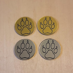 May include: Four metallic pet tag charms. Two are gold-colored, and two are silver-colored. Each charm features a black paw print design in the center. The charms are round and have a reflective surface.